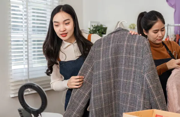 İki kadın şık bir ekose ceketi inceliyor, neşeli bir ofis ortamında çevrimiçi satış için ürünlerini dikkatlice düzenliyorlar..