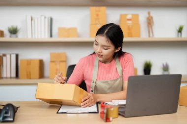 Odaklanmış bir girişimci, bir dizüstü bilgisayar üzerinde çalışırken küçük işletme operasyonlarını gösteren paketler ve notlar düzenler.