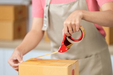 Bir kadın küçük işletmesindeki bir kutuyu kapatmak için etkili bir şekilde bant kesiciyi kullanır. Organizasyonun altını çizer ve detaylara dikkat eder..