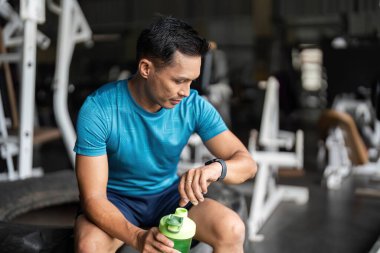 Spor kıyafetleri içinde odaklanmış bir adam, spor aletlerinin üzerine oturur, elinde protein içeceği tutarken, spor ve sağlık durumunu vurgulayarak, akıllı hareketlerini kontrol eder..