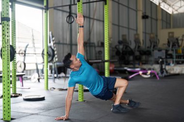 Bir adam, modern bir spor salonunda denge ve çekirdek dayanıklılığını vurgulayan bir yan tahta varyasyonu sergiliyor..