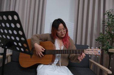 Kızıl saçlı bir kadın gitar çalıyor. O, gözlük ve kahverengi bir kazak giyiyor. Gitar kanepede.