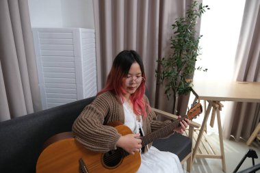 Kızıl saçlı bir kadın gitar çalıyor. O, gözlük ve kahverengi bir kazak giyiyor. Gitar kanepede.