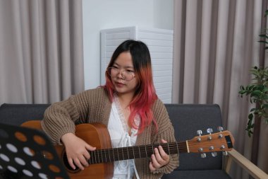 Kızıl saçlı bir kadın gitar çalıyor. O, gözlük ve kahverengi bir kazak giyiyor. Gitar kanepede.