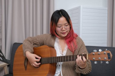 Kızıl saçlı bir kadın gitar çalıyor. O, gözlük ve kahverengi bir kazak giyiyor. Gitar kanepede.