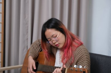 Kızıl saçlı bir kadın gitar çalıyor. O, gözlük ve kahverengi bir kazak giyiyor. Gitar kanepede.