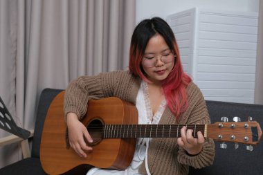 Kızıl saçlı bir kadın gitar çalıyor. O, gözlük ve kahverengi bir kazak giyiyor. Gitar kanepede.