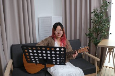 Kızıl saçlı bir kadın gitar çalıyor. O, gözlük ve kahverengi bir kazak giyiyor. Gitar kanepede.