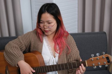 Kızıl saçlı bir kadın gitar çalıyor. O, gözlük ve kahverengi bir kazak giyiyor. Gitar kanepede.