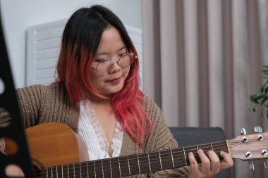 Kızıl saçlı bir kadın gitar çalıyor. O, gözlük ve kahverengi bir kazak giyiyor. Gitar kanepede.