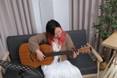 Kızıl saçlı bir kadın gitar çalıyor. O, gözlük ve kahverengi bir kazak giyiyor. Gitar kanepede.