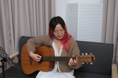 Kızıl saçlı bir kadın gitar çalıyor. O, gözlük ve kahverengi bir kazak giyiyor. Gitar kanepede.