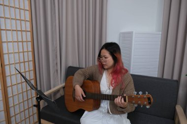 Kızıl saçlı bir kadın gitar çalıyor. O, gözlük ve kahverengi bir kazak giyiyor. Gitar kanepede.