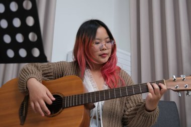 Kızıl saçlı bir kadın gitar çalıyor. O, gözlük ve kahverengi bir kazak giyiyor. Gitar kanepede.