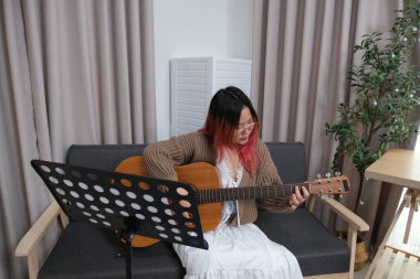 Kızıl saçlı bir kadın gitar çalıyor. O, gözlük ve kahverengi bir kazak giyiyor. Gitar kanepede.