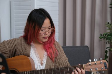 Kızıl saçlı bir kadın gitar çalıyor. O, gözlük ve kahverengi bir kazak giyiyor. Gitar kanepede.