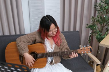 Kızıl saçlı bir kadın gitar çalıyor. O, gözlük ve kahverengi bir kazak giyiyor. Gitar kanepede.