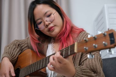 Kızıl saçlı bir kadın gitar çalıyor. O, gözlük ve kahverengi bir kazak giyiyor. Gitar kanepede.