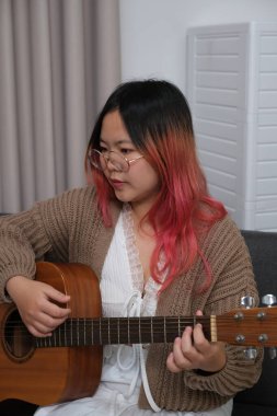 Kızıl saçlı bir kadın gitar çalıyor. O, gözlük ve kahverengi bir kazak giyiyor. Gitar kanepede.