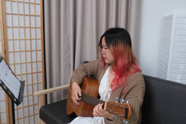 Kızıl saçlı bir kadın gitar çalıyor. O, gözlük ve kahverengi bir kazak giyiyor. Gitar kanepede.