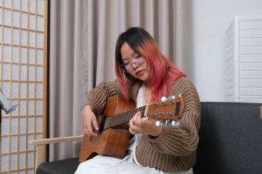 Kızıl saçlı bir kadın gitar çalıyor. O, gözlük ve kahverengi bir kazak giyiyor. Gitar kanepede.