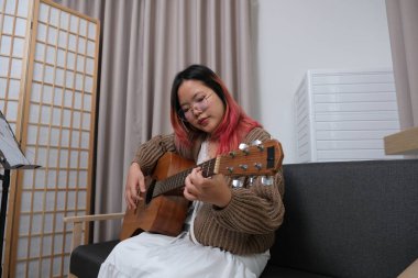 Kızıl saçlı bir kadın gitar çalıyor. O, gözlük ve kahverengi bir kazak giyiyor. Gitar kanepede.