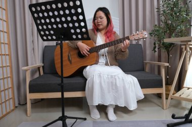 Kızıl saçlı bir kadın gitar çalıyor. O, gözlük ve kahverengi bir kazak giyiyor. Gitar kanepede.