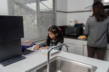 Annesi kahvaltı hazırlarken, aile etkileşimini vurgularken ve öğrenirken mutfak masasında derslerine odaklanan genç bir kız..