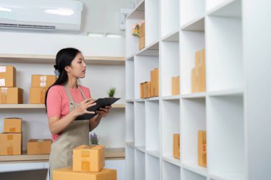 Genç bir kadın, küçük işletme verimliliğini vurgulayarak envanterini iyi organize edilmiş bir ev ofisinde düzenlemek için bir tablet kullanıyor..