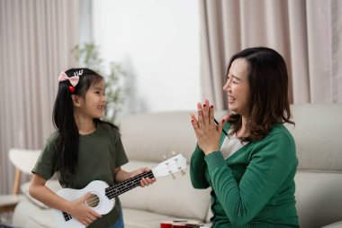 Bir anne, kızı ukulele çalarken alkışlar ve gülümser. Müziğin ve aile bağının neşesini vurgular..