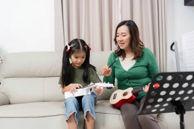 Bir anne kızına ukulele çalma konusunda rehberlik eder, sıcak bir evde müzik eğitimi ve aile bağlarını teşvik eder..