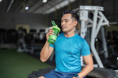 Formda bir adam protein karıştırıcısından bir yudum alır ve fitness rutinlerinde hidrasyonun önemini vurgular..