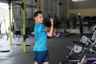 Spor salonunda kolunu uzatan odaklanmış bir adam antrenmana hazırlanıyor. Zindelik ve esnekliği vurgular.