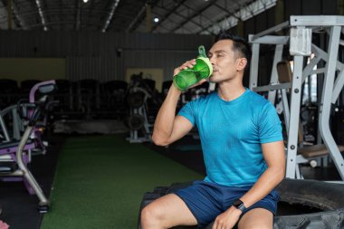 Formda bir adam protein karıştırıcısından ferahlatıcı bir yudum alır. Egzersiz sırasında susuzluğun önemini vurgular..