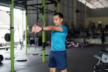 Genç bir adam spor salonunda kol esnetme hareketleri yapıyor, esnekliği ve antrenmanlara hazırlanmayı vurguluyor..