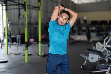 Odaklanmış bir adam spor salonunda esnemek için koluna uzanıyor. Fitness ve esnekliği vurguluyor. Sağlık ve refahı ön plana çıkarıyor.