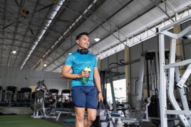 Elinde su şişesi ve spor çantası olan gülümseyen bir adam geniş bir spor salonunda yürüyor. Fitness yaşam tarzı ve pozitifliğe vurgu.