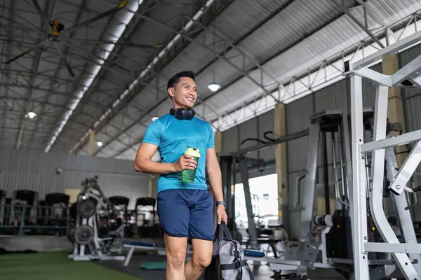 Elinde su şişesi ve spor çantası olan gülümseyen bir adam geniş bir spor salonunda yürüyor. Fitness yaşam tarzı ve pozitifliğe vurgu.