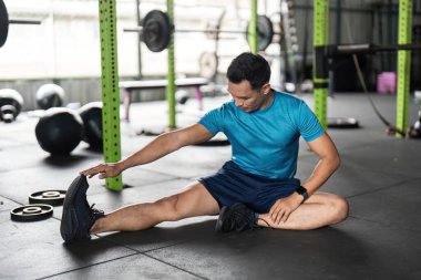 Mavi tişörtlü kendini işine adamış bir adam spor salonunda ayak bileğini esnetiyor. Spor yaparken spor yaparken vücut geliştirme ve esnekliğini ön plana çıkarıyor..