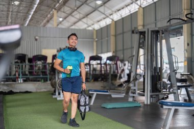 Neşeli bir adam elinde protein içeceği ve spor çantasıyla spor salonuna giriyor. Pozitif fitness yaşam tarzını ve motivasyonunu temsil ediyor..