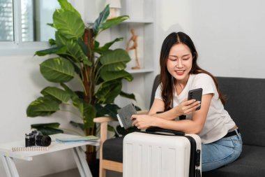 Kanepede oturan bir kadın, akıllı telefonunu kontrol ederken yaklaşan bir yolculuğa hazırlanıyor, seyahat hazırlığını vurguluyor..