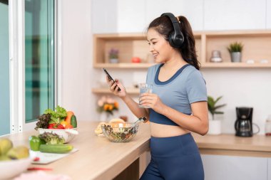 Neşeli genç bir kadın bir bardak su tutuyor ve sağlıklı yaşam ve sürdürülebilirliği vurgulayarak taze salata yaparken telefonunu kontrol ediyor..