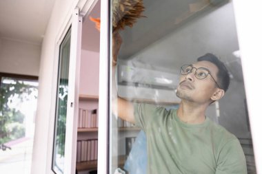 Pencereyi temizlemeye odaklanan bir adam, detaylara dikkat eder ve güneşli bir ortamda ev bakımına bağlılığını gösterir..