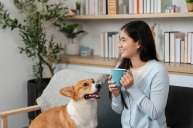 Genç bir kadın elinde bir fincan kahve tutarken gülümsüyor, Corgi köpeği dikkatle ona bakıyor, evinde samimi bir anı yakalıyor..