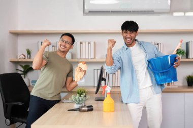 İki erkek ofis temizliği görevlerini tamamladıktan sonra sevinç ve memnuniyet ifade ediyor, takım çalışmasını ön plana çıkarıyor.