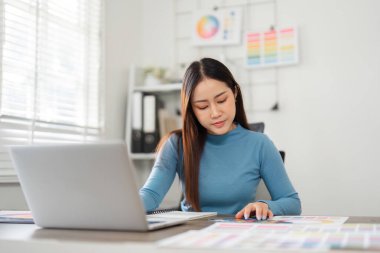 Bir kadın tasarımcı, parlak çalışma alanında renk örneklerini, beyin fırtınalarını ve yaratıcı girişimlerini incelikle karşılaştırır..