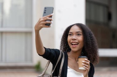 Bir anı paylaşmak için heyecanlanan genç kadın, elinde kahve fincanıyla parlak bir selfie çekerken, canlı şehir yaşam tarzını sergiliyor..