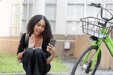 Genç bir kadın, şehir parkında yeşil bisikletinin yanında oturmuş, doğanın ortasında modern bağlantıyı kucaklayan bir video görüşmesinden hoşlanıyor..