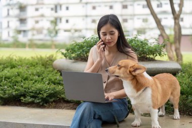 Corgi köpeği yanında oturup uzaktan çalışmanın ve evcil hayvan arkadaşlığının zevklerini vurgularken bir kadın telefonuyla iş ve iletişimi dengeliyor..