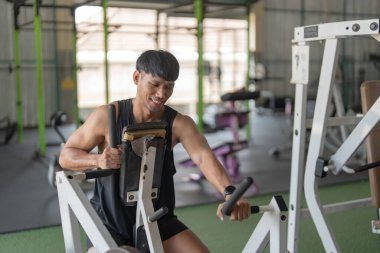 Bir adam spor makinesi kullanırken gülümser, güç antrenmanı ve spor yapma motivasyonunu vurgular..